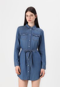 Robe en denim avec un col, des manches longues et deux poches poitrine. Elle possède une taille ceinturée et un ourlet arrondi. Ton bleu clair.