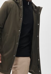 Man som bär en olivgrön jacka med dragkedja över en svart kabelstickad tröja och beige byxor framför en vit bakgrund.