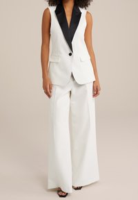 Ärmlös vit blazer med svarta slag och en enda knapp, i kombination med vida vita byxor och svarta klacksandaler.