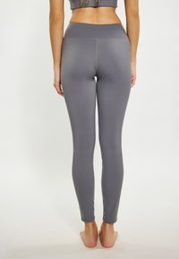 Szürke leggings sima, rugalmas anyagból, testhezálló fazonban és széles derékrésszel, hátulról nézve, mezítláb egy sima háttér előtt.