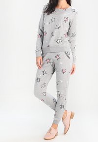 Eine Frau steht da und trägt einen grauen Jogginganzug mit passenden Sweatshirt und Jogginghosen, die mit pinken und schwarzen Sternmustern versehen sind, kombiniert mit hellpinkfarbigen Oxfordschuhen.