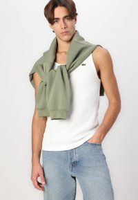 Hvit ribbet tank topp med en olivengrønn hettegenser drapert over skulderne. Lette blå denimjeans. Lacoste-logo på tanken.