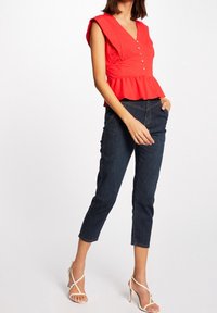 Röd peplumtop med V-ringning och knapprader, kombinerad med mörk denimcapribyxor och vita sandaler med remmar.