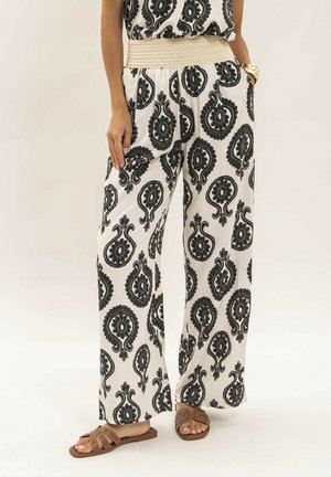 Femme portant un pantalon blanc à jambes larges avec de grands motifs cachemire noirs, une ceinture tissée beige et des sandales ouvertes marron.