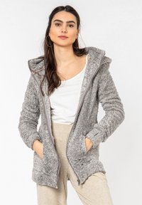 Gray fleece zip-up jacket with a hood, featuring side pockets and ribbed cuffs. Worn over a white top and beige bottoms. Soft texture.