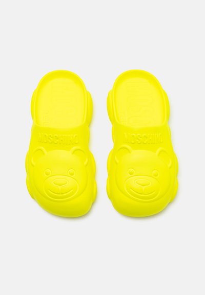Chanclas de goma amarillas con un diseño de cara de oso en la parte superior, con la palabra 'MOSCHINO' grabada en cada zapato.
