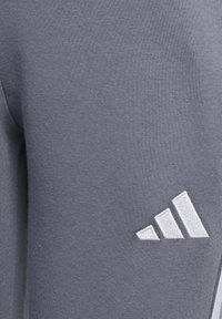 Graue Sporthose aus weichem Stoff mit einem gestickten weißen Drei-Streifen-Logo auf der Außenseite. Glatte Textur ohne Muster.