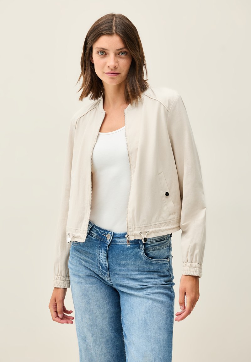 Ljust beige bomberjacka med dragkedja fram, elastiska muddar och dragsko i nederkant. Bärs över en vit tanktop och blå jeans.