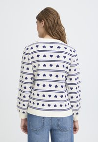 Pull en mélange de coton avec une base crème ornée de motifs de cœurs bleu marine et de rayures horizontales. Les poignets et l'ourlet côtelés apportent de la texture.