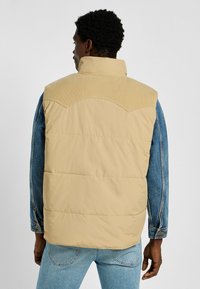 Beige pufferväst med en kordfloddyok, tillsammans med en denimjacka. Vesten har en fyrkantig form och textil med struktur i den övre delen.