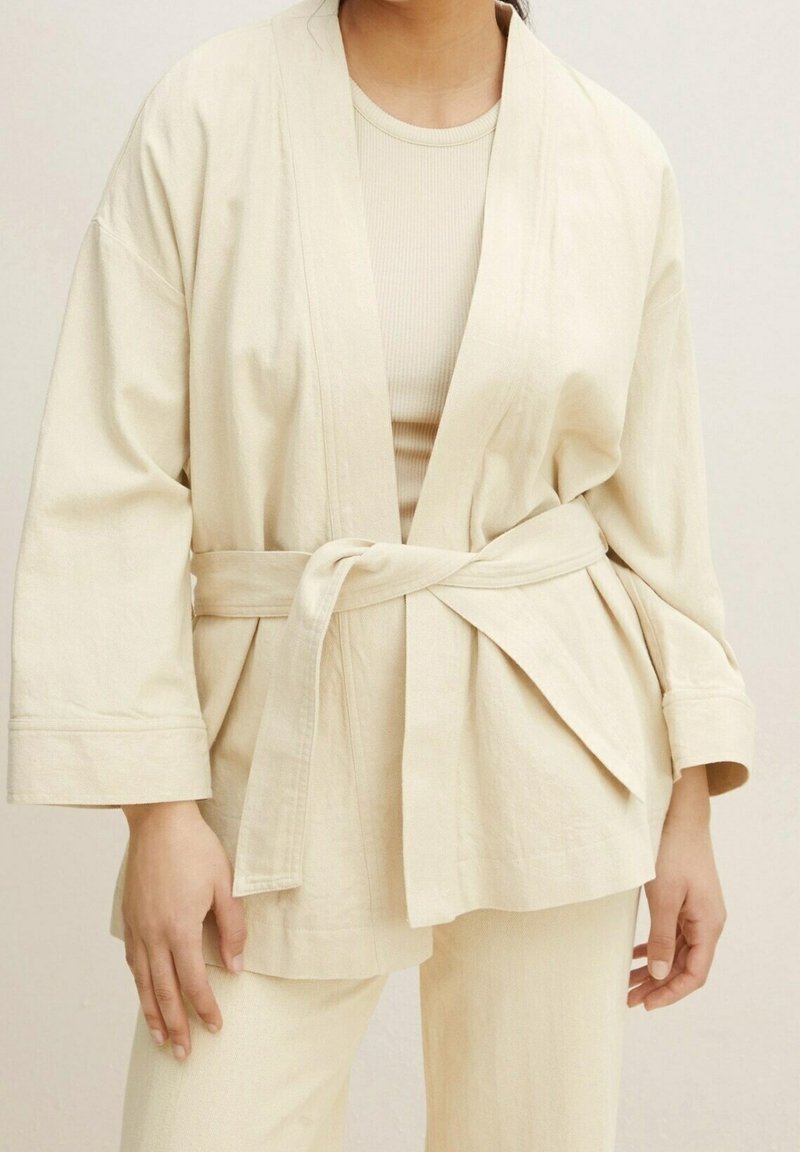 Femme portant une veste kimono écrue ceinturée sur un haut côtelé et un pantalon assorti, debout dans une posture détendue devant un fond neutre.