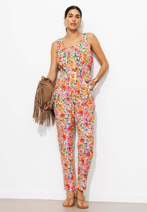 Femme en combinaison fleurie sans manches avec taille à cordon, tenant un sac marron à franges, portant des sandales et des boucles d’oreilles créoles dorées, debout contre un mur blanc.