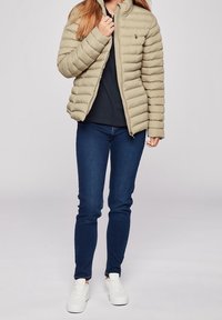 Beige puffy jacket with a zip closure and high collar, paired with dark blue jeans and white sneakers in a front view.