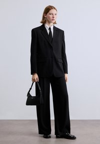 Μαύρο σμόκιν με ευρείς παντελόνια και εφαρμοστή σακάκι, σετ με λευκό πουκάμισο και μαύρη γραβάτα. Τα αξεσουάρ περιλαμβάνουν μια μικρή μαύρη τσάντα.