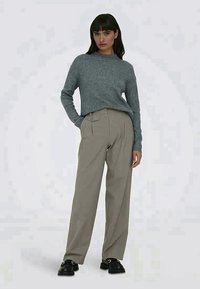 Γυναίκα που στέκεται φορώντας ένα γκρι ριμπ εσώρουχο, μπέζ ψηλόμεσες πλατιά παντελόνες και μαύρους λοφούς, ποζάροντας μπροστά σε μια απλή φόντο.