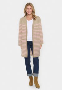 Beige kofta med ljusrosa ränder, öppen front och fickor. Bärs över en vit topp med mörka jeans och bruna, klackade anklarstövlar.