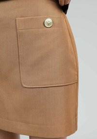 Jupe beige en tissu texturé, dotée d'une poche avant, de coutures décoratives et d'un accent avec un bouton doré au design de logo.
