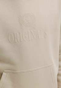 Sweat-shirt beige clair avec texte embossé "ORIGINALS" et un emblème rond avec une couronne de laurier et un texte plus petit au-dessus de la poche.