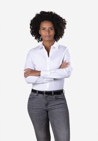 Confident woman with natural curly hair, wearing a white button-up shirt and gray jeans, stands with arms crossed against a plain background.