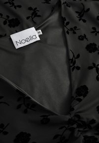 Čierna kvetinová látka s hladkým, mierne lesklým povrchom. Má výstrih do v a nariasenie v strede. Viditeľný štítok značky.
