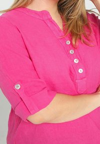 Chemise en lin fuchsia avec col mandarin, manches trois-quarts et patte de boutonnage. Comprend une petite poche à l'avant.
