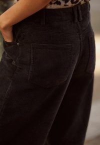 Pantalons en velours côtelé noir avec des côtes verticales, dotés d'une poche arrière unique et d'une coupe décontractée. La texture semble douce et légèrement veloutée.
