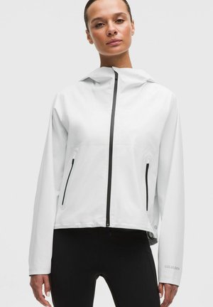 Vrouw die een wit jack met ritssluiting draagt, met zwarte details aan de rits, en zwarte broek, staand tegen een eenvoudige lichte achtergrond.