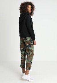 Felpa con cappuccio nera abbinata a pantaloni mimetici verdi con motivi floreali neri, completata da sneakers bianche.