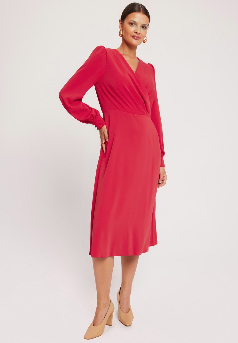 Czerwona sukienka z długim rękawem w stylu kopertowym i dekoltem w kształcie litery V, o gładkiej fakturze i długości do kolan, zestawiona z beżowymi szpilkami.