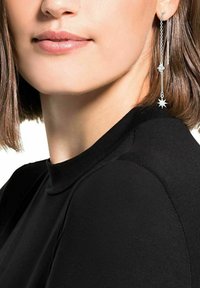 Kvinna med svart topp och kort brunt hår, sedd från hakan till axeln, med en lång silverörhänge med stjärncharms.