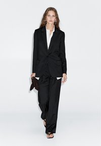 Schwarz maßgeschneiderter Anzug mit einem taillierten Blazer und weiten Hosen, über einem weißen Hemd getragen. Die Accessoires umfassen eine dunkle Clutch und Sandalen.