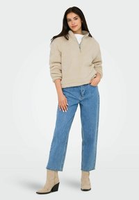 Pull en polaire beige avec un col zippé d'un quart et une texture, associé à un jean en denim bleu taille haute et des bottines beiges.