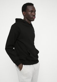 Svart hettegenser med frontlomme, laget av myk stoff, med et enkelt design og brodert logo. Kombinert med lysegrå joggebukser.