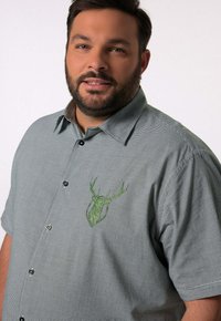 Camisa de botones de manga corta en un patrón de cuadros en verde y blanco, que cuenta con un bordado de una cabeza de ciervo verde en el área del pecho izquierdo.