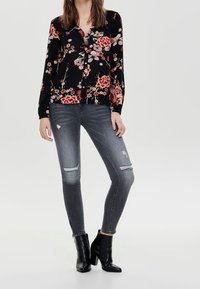 Mujer vestida con una blusa floral negra con botones, jeans ajustados rasgados grises y botines de tacón negros, de pie contra un fondo liso.