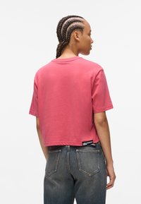 Skraćeni, majica s kratkim rukavima u dusty pink pamuku s okruglim izrezom. Tag na leđima sadrži oznaku "Karl Lagerfeld Jeans". Nosena s tamnim trapericama.