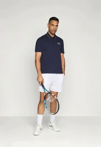 Σκούρο μπλε πόλο μπλούζα με γιακά και λευκή επένδυση, με λογότυπο. Φορεμένη με λευκά σορτς, κρατώντας μια ρακέτα τένις, στέκεται σε απλό φόντο.