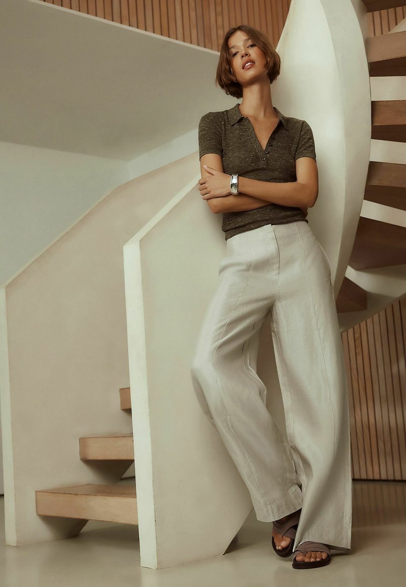 Donna con capelli corti che indossa una polo scura, pantaloni chiari a gamba larga e sandali, appoggiata a una scala a chiocciola bianca all'interno di un edificio.