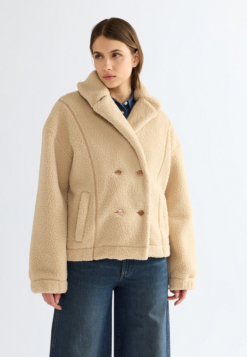 Femme portant une veste en laine beige à double boutonnage avec un large col et un jean bleu en denim, debout devant un fond blanc uni.