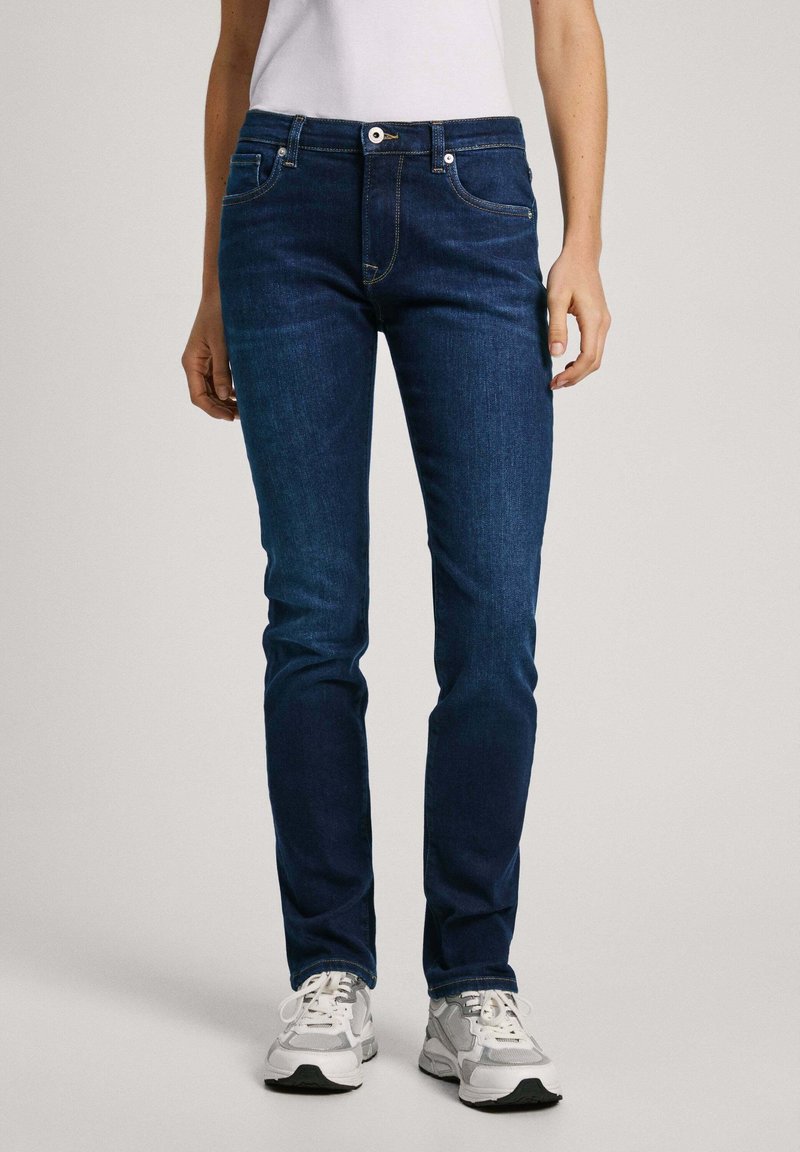 Mörkblå jeans av denim, slim fit, med framfickor och en enkel knappstängning. Kombinerade med gråa sportskor.