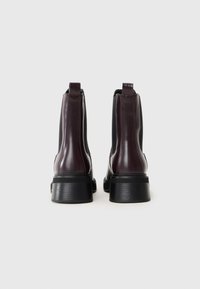 Botines de cuero burdeos con tacones de goma negra, que cuentan con inserciones elásticas en los laterales y tiradores para facilitar su uso. Textura suave con acentos cosidos.