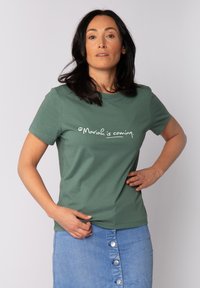 T-shirt en coton vert à manches courtes avec le texte blanc "Mariah is coming." Femme portant une jupe en denim avec des boutons.