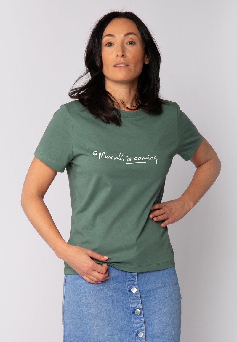 T-shirt en coton vert à manches courtes avec le texte blanc "Mariah is coming." Femme portant une jupe en denim avec des boutons.