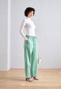 White long-sleeve fitted top paired with loose-fitting mint green pants with an elastic waistband and side pockets; model holding a small bag.