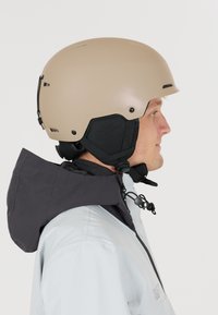 Man met een beige helm met zwarte kinband en een zwarte winterjack met verhoogde capuchon, die naar rechts kijkt tegen een effen witte achtergrond.