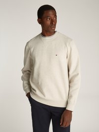 Sweatshirt beige claro com gola redonda e punhos canelados. Apresenta um pequeno logótipo no peito. Usado com calças de cor azul-marinho sobre um fundo neutro.