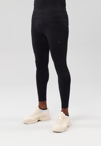 Leggings pretos feitos de um tecido elástico, com uma textura suave e um pequeno detalhe de logotipo. Combinados com sapatilhas de desporto de cor clara.