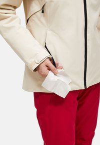 Håndtrækker et hvidt væv ud af en lynlåslomme på en beige jakke, der er iført med røde bukser, isoleret på en ensfarvet baggrund.