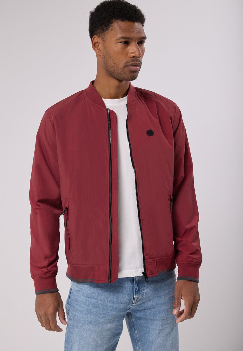 Veste bomber légère rouge avec des poignets côtelés, fermeture éclair, poches latérales et un détail logo noir. Portée par-dessus un t-shirt blanc.