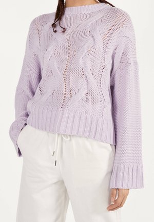 Person wearing a lavender cable-knit sweater with ribbed cuffs and hem, paired with white drawstring pants, hands relaxed at sides.